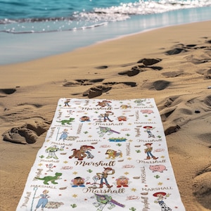 May include: A white beach towel with a cartoon design featuring characters and the name "Marshall" repeated throughout. The towel is lying on a sandy beach with the ocean in the background.