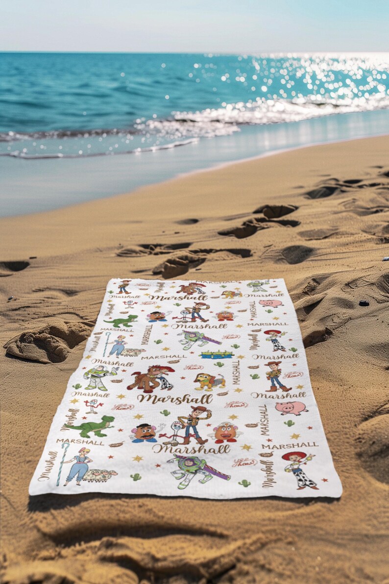 May include: A white beach towel with a cartoon design featuring characters and the name "Marshall" repeated throughout. The towel is lying on a sandy beach with the ocean in the background.