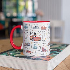 May include: A white ceramic mug with a red interior and handle, decorated with cartoon cars and the name "Jordi". The design includes various car illustrations, stars, and the name "Jordi" repeated across the surface.