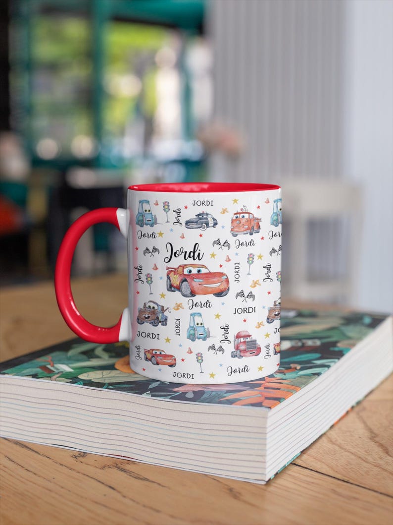 May include: A white ceramic mug with a red interior and handle, decorated with cartoon cars and the name "Jordi". The design includes various car illustrations, stars, and the name "Jordi" repeated across the surface.