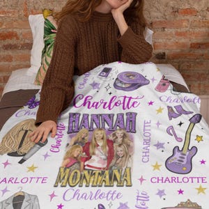 Op de afbeelding: Een witte deken met een ontwerp met de tekst "Charlotte" en "Hannah Montana". De deken bevat afbeeldingen van een gitaar, een tv, een vlinder en een cd. De deken is versierd met sterren en andere afbeeldingen.