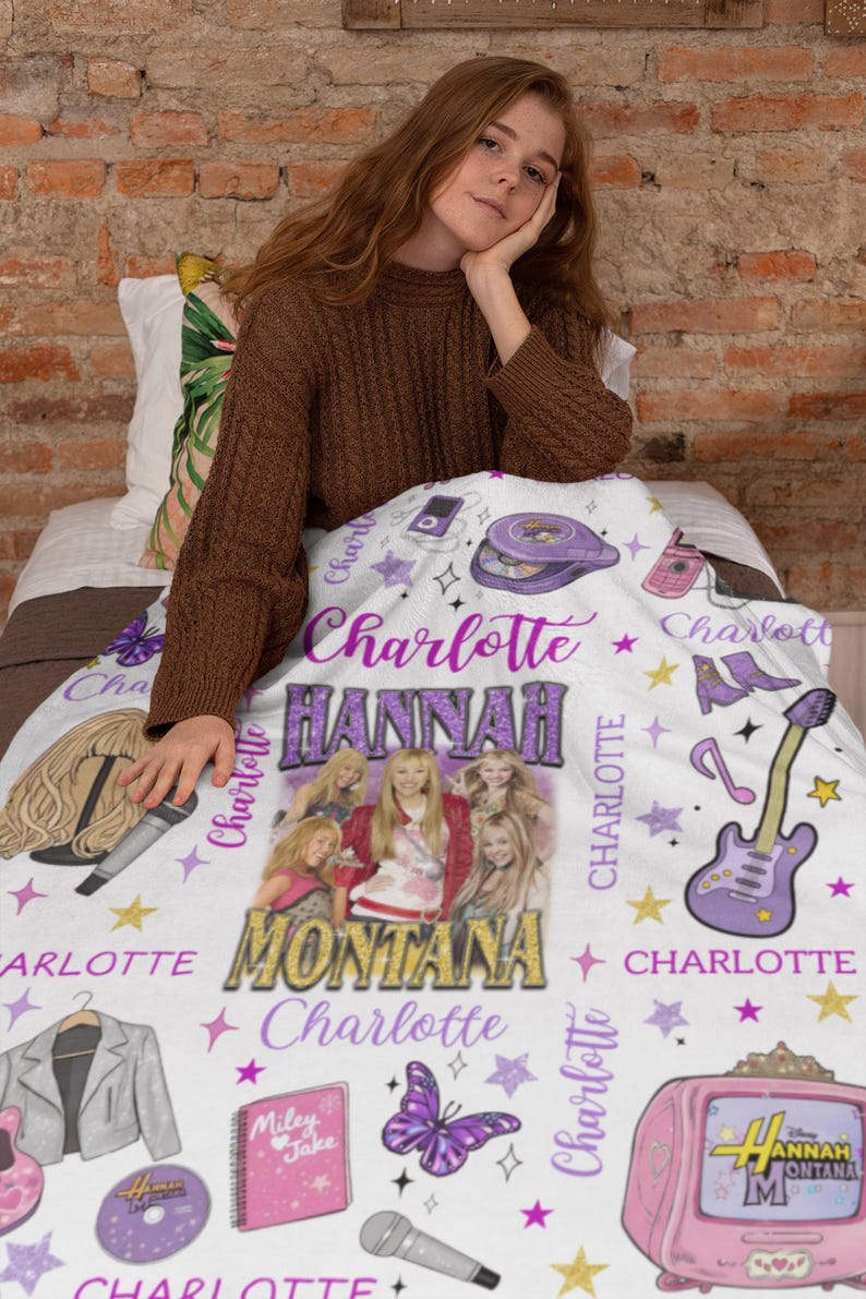Op de afbeelding: Een witte deken met een ontwerp met de tekst "Charlotte" en "Hannah Montana". De deken bevat afbeeldingen van een gitaar, een tv, een vlinder en een cd. De deken is versierd met sterren en andere afbeeldingen.