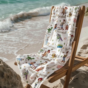 May include: White beach towel with a repeating pattern of cartoon characters and the name "Marshall". The towel is draped over a wooden beach chair on a sandy beach, with the sea visible in the background. The design is colourful and playful.