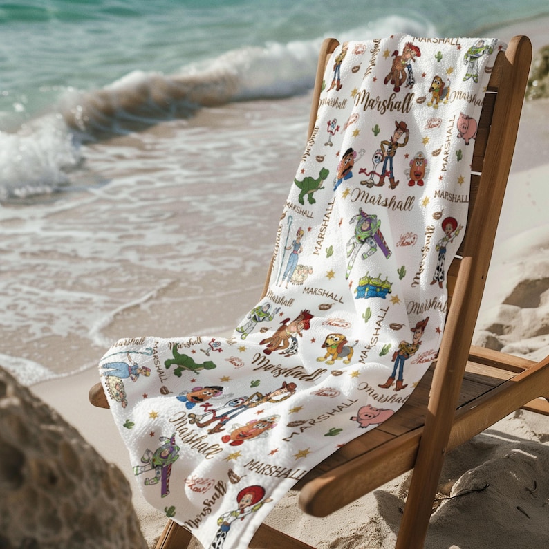 May include: White beach towel with a repeating pattern of cartoon characters and the name "Marshall". The towel is draped over a wooden beach chair on a sandy beach, with the sea visible in the background. The design is colourful and playful.