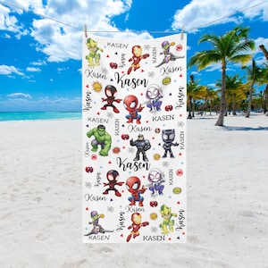 Pu&ograve; includere: Telo mare bianco con personaggi di supereroi dei cartoni animati colorati e il nome "Kasen" ripetuto. L'asciugamano &egrave; appeso a un filo, con una spiaggia e un cielo blu sullo sfondo.