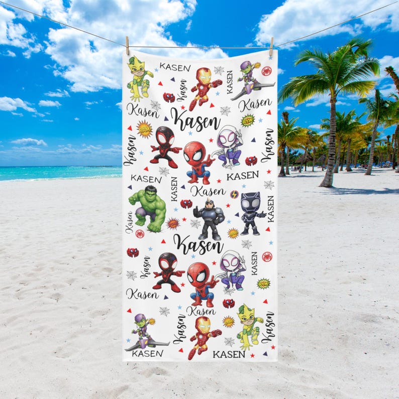 Pu&ograve; includere: Telo mare bianco con personaggi di supereroi dei cartoni animati colorati e il nome "Kasen" ripetuto. L'asciugamano &egrave; appeso a un filo, con una spiaggia e un cielo blu sullo sfondo.