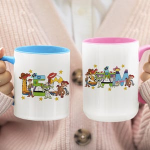 Puede incluir: Dos tazas de cer&aacute;mica blanca con dise&ntilde;os coloridos. Una taza tiene el interior y el asa azules, la otra tiene el interior y el asa rosas. Las tazas presentan la palabra "TEAM" con personajes de dibujos animados y estrellas.