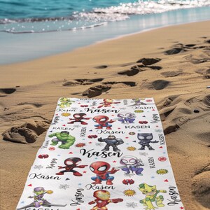 Pu&ograve; includere: Un telo da mare bianco con personaggi di supereroi dei cartoni animati colorati e il nome "Kasen" in un font stilizzato. L'asciugamano &egrave; decorato con stelle ed elementi in stile fumetto. L'asciugamano &egrave; su una spiaggia sabbiosa con l'oceano sullo sfondo.