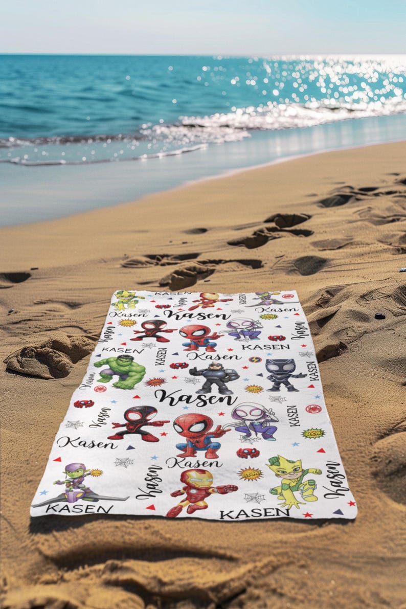 Pu&ograve; includere: Un telo da mare bianco con personaggi di supereroi dei cartoni animati colorati e il nome "Kasen" in un font stilizzato. L'asciugamano &egrave; decorato con stelle ed elementi in stile fumetto. L'asciugamano &egrave; su una spiaggia sabbiosa con l'oceano sullo sfondo.