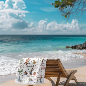 May include: A white beach towel featuring cartoon characters and the name "Marshall". The towel is on a wooden beach chair on a sandy beach. The background shows a blue sea and sky with clouds.