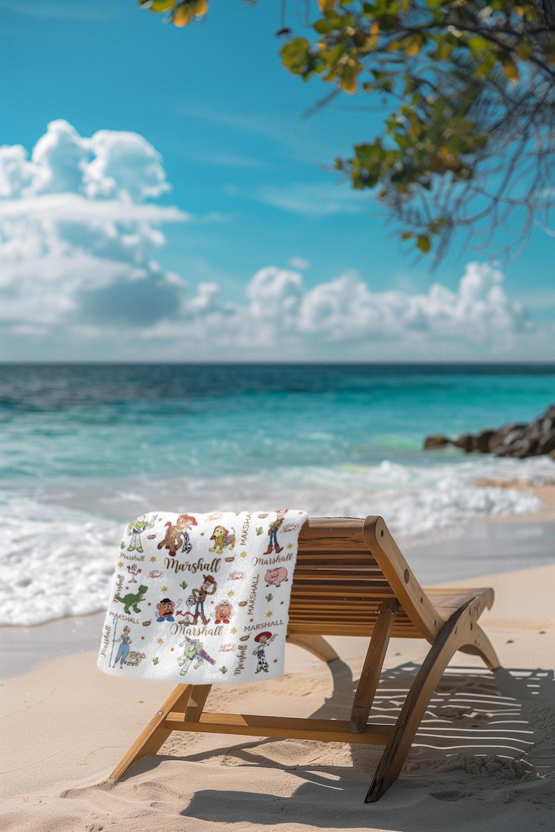 May include: A white beach towel featuring cartoon characters and the name "Marshall". The towel is on a wooden beach chair on a sandy beach. The background shows a blue sea and sky with clouds.