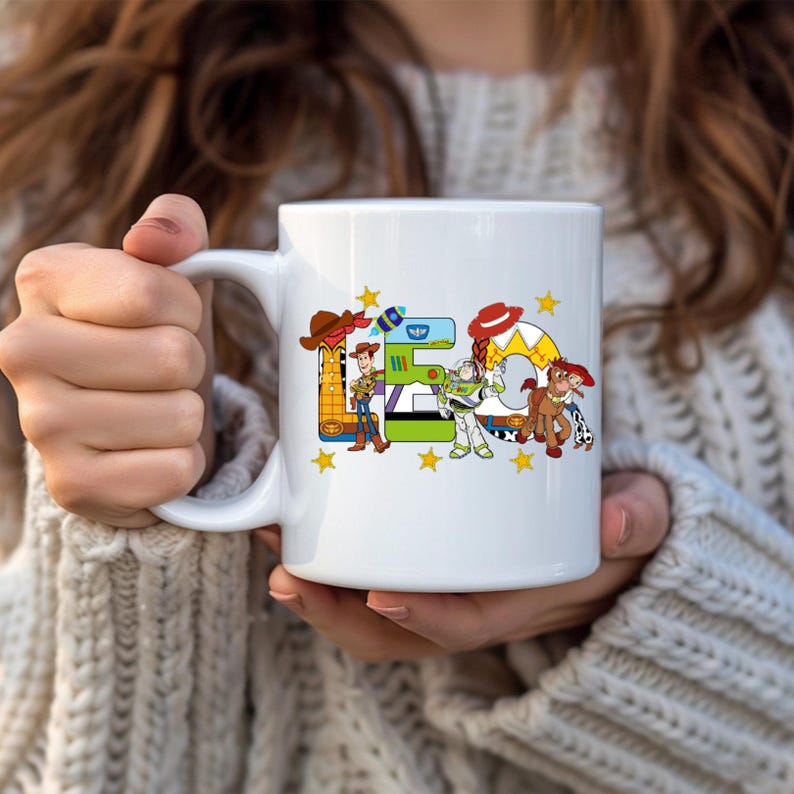 Puede incluir: Taza de cer&aacute;mica blanca con la palabra "E" y un sombrero de vaquero, Woody y un cohete. El n&uacute;mero "5" es verde, y la letra "G" tiene un sombrero rojo y un caballo. Tambi&eacute;n aparece Buzz Lightyear. Estrellas amarillas est&aacute;n dispersas alrededor del dise&ntilde;o.