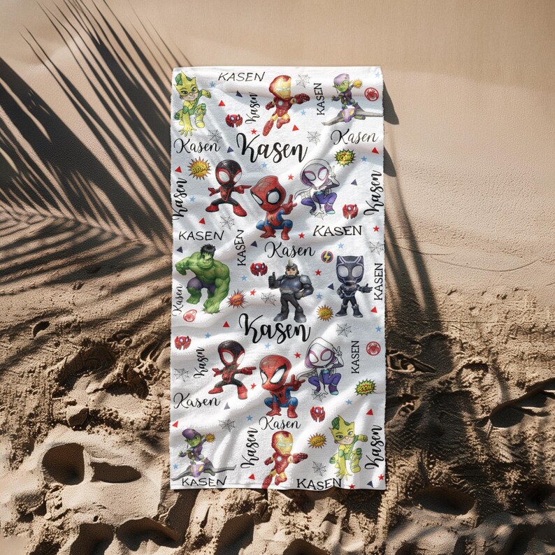 Pu&ograve; includere: Telo mare bianco con illustrazioni di supereroi dei cartoni animati e il nome "Kasen" scritto a mano. L'asciugamano presenta un motivo ripetuto di supereroi, stelle e raggi di sole. L'asciugamano &egrave; appoggiato su una superficie sabbiosa.