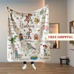 May include: A cream-colored blanket featuring various Toy Story characters, including Woody, Buzz Lightyear, and others. The name "Hudson" is repeated throughout the design, along with stars and cacti. The blanket is held up by a person.