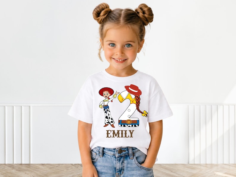 Puede incluir: Una camiseta blanca con una ilustración de dibujos animados de Jessie de Toy Story, el número 2 y el nombre "EMILY". El diseño incluye un sombrero rojo, pelo amarillo y un estampado de vaca. La niña lleva vaqueros azules.