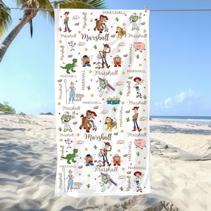 May include: White beach towel with a repeating pattern of cartoon characters, stars, and cacti. The towel has the word "Marshall" in a gold script font. The towel is hanging on a clothesline at the beach.