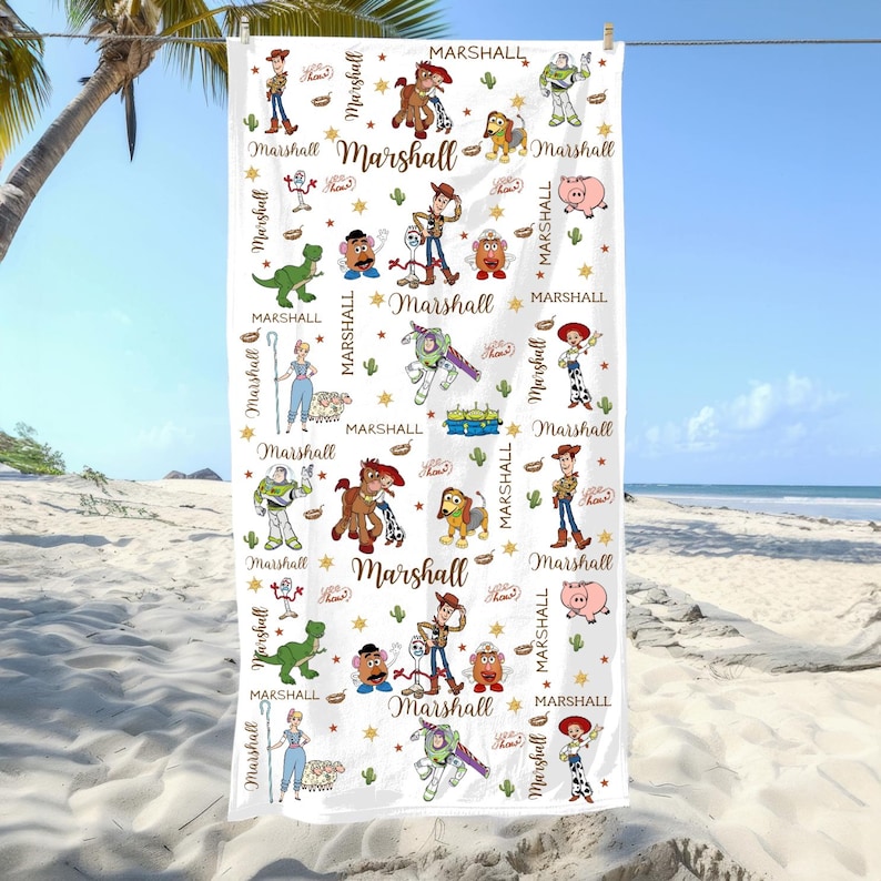 May include: White beach towel with a repeating pattern of cartoon characters, stars, and cacti. The towel has the word "Marshall" in a gold script font. The towel is hanging on a clothesline at the beach.