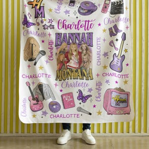 Op de afbeelding: Witte deken met een ontwerp met de tekst "Hannah Montana" en "Charlotte". Het ontwerp bevat afbeeldingen van een gitaar, microfoon, vlinder en andere muzikale elementen. De deken wordt door een persoon vastgehouden.