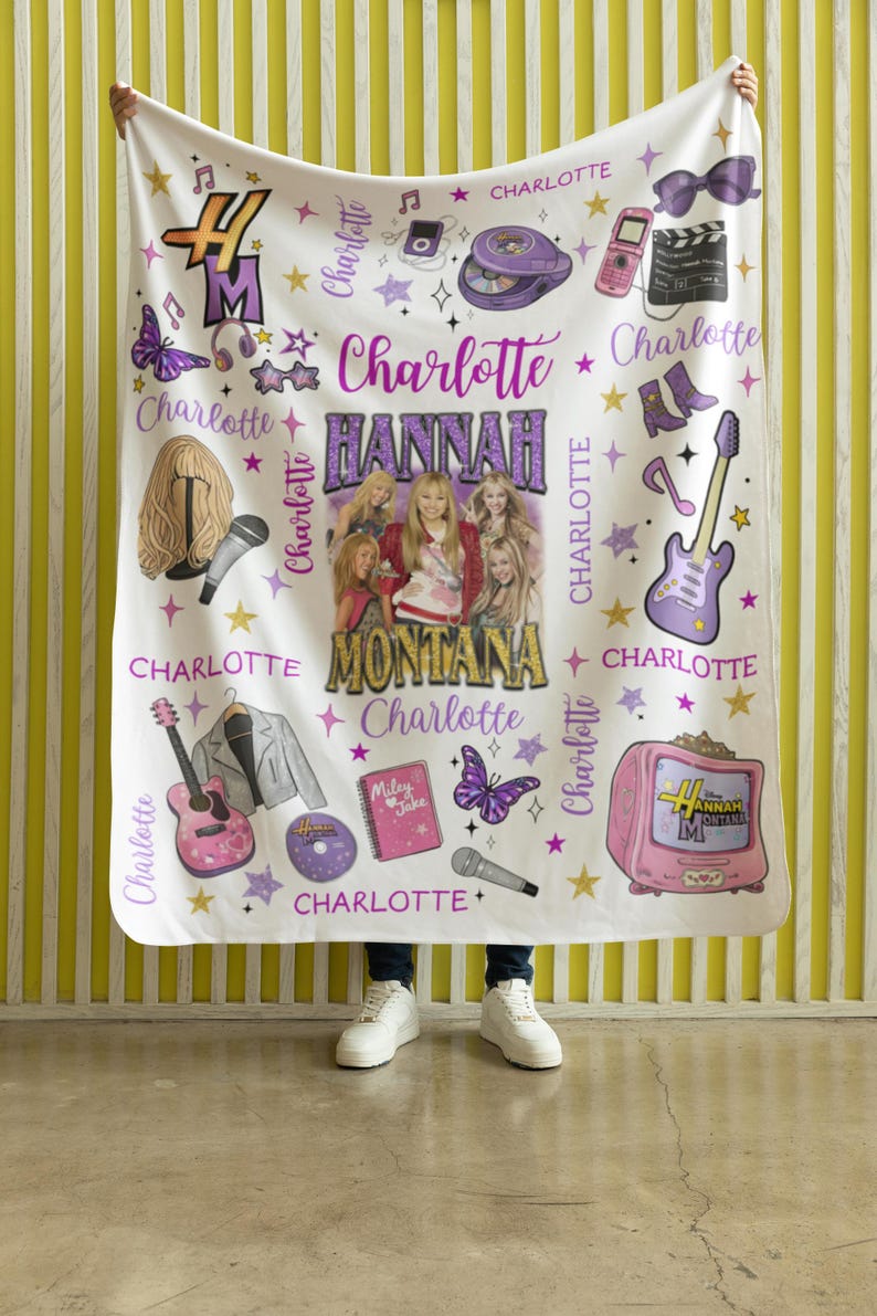 Op de afbeelding: Witte deken met een ontwerp met de tekst "Hannah Montana" en "Charlotte". Het ontwerp bevat afbeeldingen van een gitaar, microfoon, vlinder en andere muzikale elementen. De deken wordt door een persoon vastgehouden.