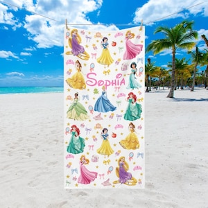 May include: White beach towel featuring various cartoon princesses in colorful gowns. The towel has the name "Sophia" in pink script. Decorative elements include crowns, bows, and apples. The towel is hanging on a clothesline against a beach backdrop.