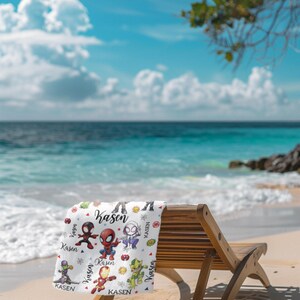Pu&ograve; includere: Un telo mare bianco con personaggi di supereroi dei cartoni animati e il nome "Kasen" stampato. L'asciugamano &egrave; appoggiato su una sedia a sdraio di legno su una spiaggia sabbiosa, con l'oceano e il cielo blu sullo sfondo.