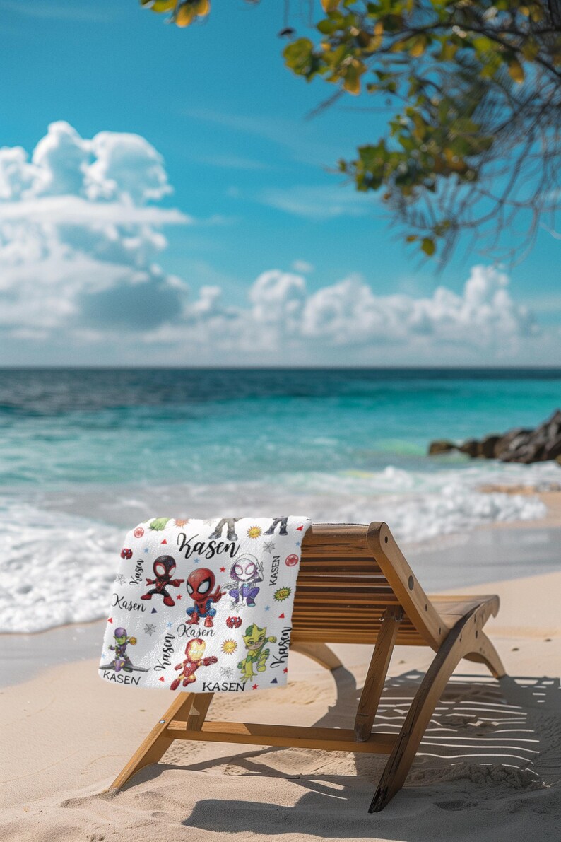 Pu&ograve; includere: Un telo mare bianco con personaggi di supereroi dei cartoni animati e il nome "Kasen" stampato. L'asciugamano &egrave; appoggiato su una sedia a sdraio di legno su una spiaggia sabbiosa, con l'oceano e il cielo blu sullo sfondo.