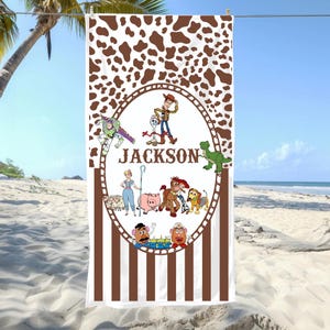 May include: A white beach towel with a brown cow-print top and vertical brown and white stripes at the bottom. The center features a cartoon design with the name "JACKSON" and various character illustrations.