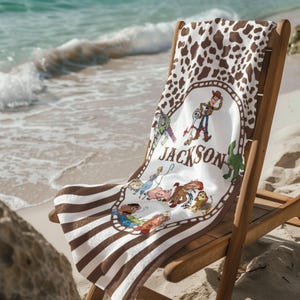 May include: A beach towel featuring a brown and white cow print pattern at the top and brown and white stripes at the bottom. The center has a white oval with cartoon characters and the name "Jackson". The towel is draped over a wooden beach chair.