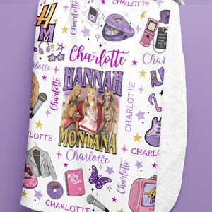Op de afbeelding: Witte fleece deken met een ontwerp met de tekst "Hannah Montana" en "Charlotte" in paars en goud. De deken bevat afbeeldingen van een microfoon, sterren, een vlinder en een roze televisie.