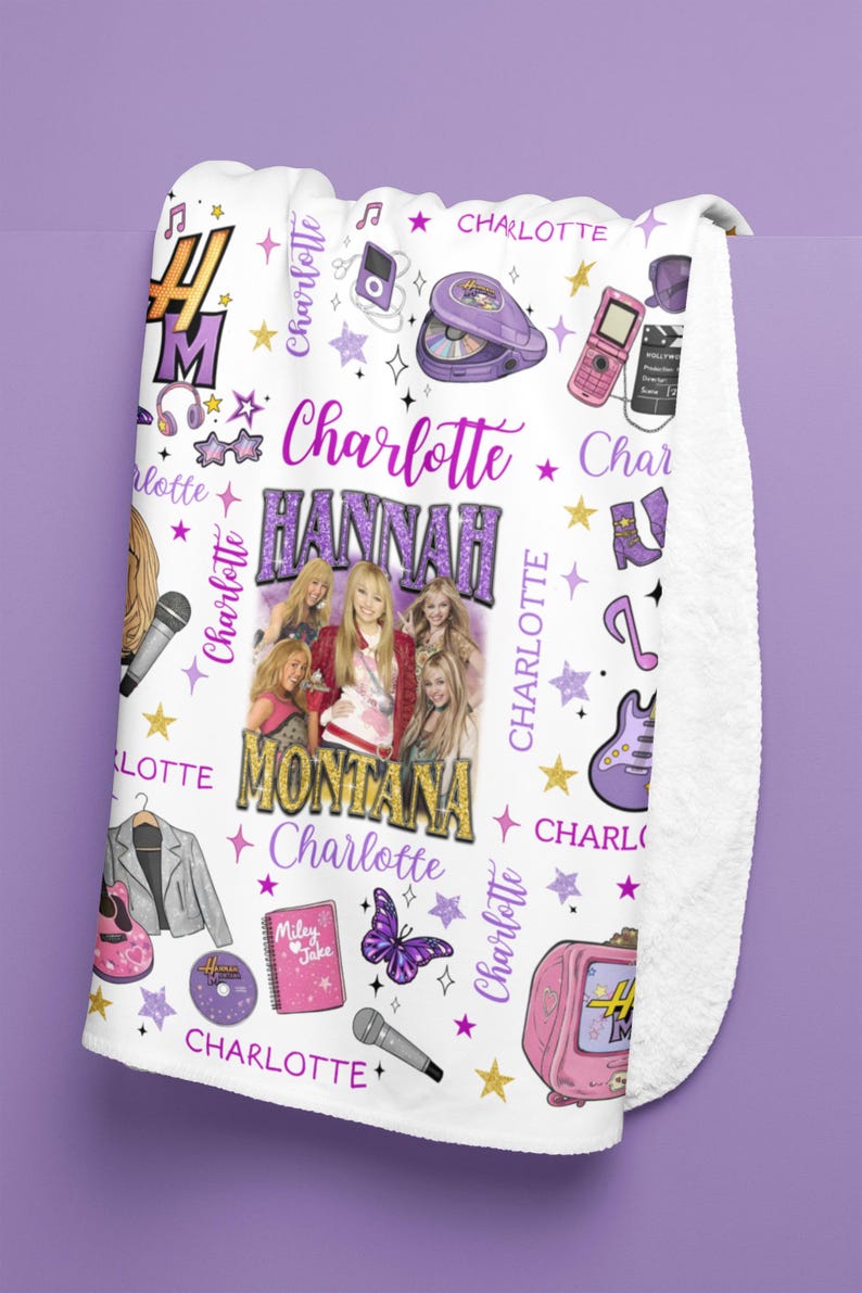 Op de afbeelding: Witte fleece deken met een ontwerp met de tekst "Hannah Montana" en "Charlotte" in paars en goud. De deken bevat afbeeldingen van een microfoon, sterren, een vlinder en een roze televisie.