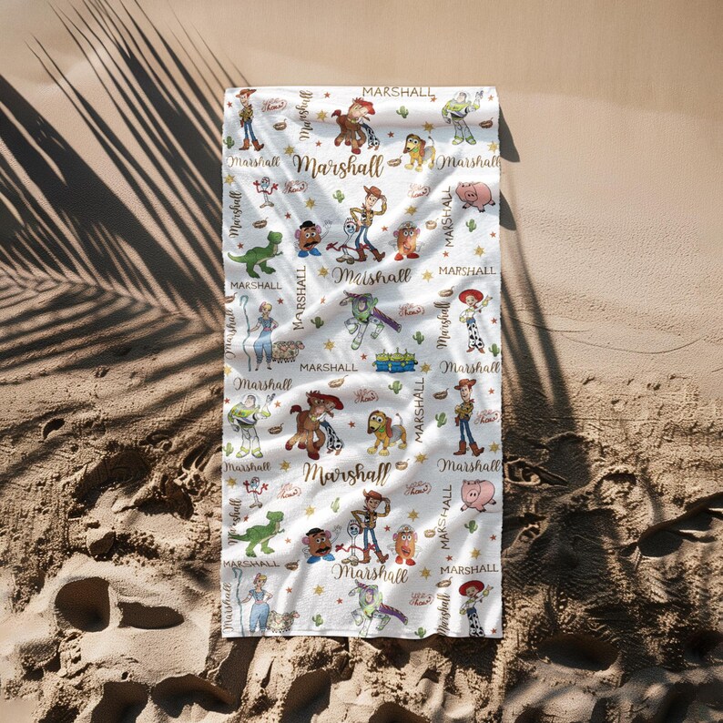 May include: White beach towel with a repeating pattern of cartoon characters and the name "Marshall" in gold script. The towel features images of a cowboy, a space ranger, a pig, and other characters.