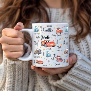 May include: A white ceramic mug with a black handle and the text "But First, Coffee" in a black, handwritten-style font. The mug is on a white surface.