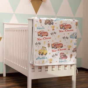 Puede incluir: Una cuna blanca con una manta con dibujos de coches de dibujos animados, rayos y el texto "Ka-Chow" y "Hollis". La manta tiene un patr&oacute;n repetido de coches de carreras, trofeos y banderas a cuadros.