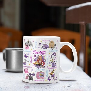 K&ouml;nnte beinhalten: Wei&szlig;e Keramik-Tasse mit einem farbenfrohen Design mit dem Namen "Charlotte" und Illustrationen von Gitarre, Mikrofon und Fernseher. Die Tasse hat einen gro&szlig;en Henkel und ist mit einer dunklen Fl&uuml;ssigkeit gef&uuml;llt. Im Hintergrund befindet sich eine silberne Tasse.
