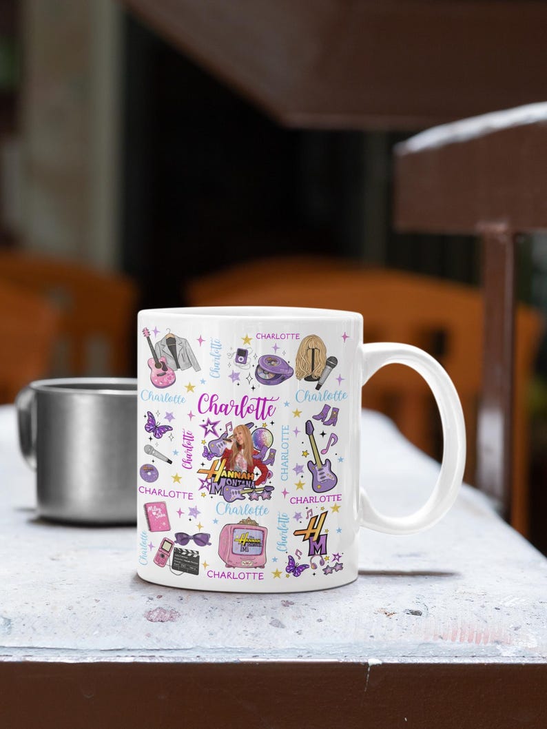 K&ouml;nnte beinhalten: Wei&szlig;e Keramik-Tasse mit einem farbenfrohen Design mit dem Namen "Charlotte" und Illustrationen von Gitarre, Mikrofon und Fernseher. Die Tasse hat einen gro&szlig;en Henkel und ist mit einer dunklen Fl&uuml;ssigkeit gef&uuml;llt. Im Hintergrund befindet sich eine silberne Tasse.