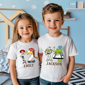 以下が含まれることがあります： 白いTシャツを着た2人の子供たち。1つのシャツには、数字の2と「Emily」という名前が付いたウッディとジェシーのデザインが施されています。もう1つのシャツには、数字の4と「Jackson」という名前が付いたバズ・ライトイヤーのデザインが施されています。