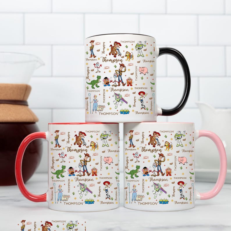 Puede incluir: Tres tazas de cer&aacute;mica blanca con dise&ntilde;os de personajes de dibujos animados coloridos y el nombre "Thompson" impreso. Una taza tiene un asa negra, otra roja y otra rosa. Las tazas est&aacute;n apiladas.