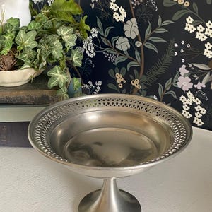 May include: A silver-colored pedestal serving dish with a decorative pierced rim. The dish has a round bowl shape and a short, rounded pedestal base. The background features a floral wallpaper and a small white bird figurine.