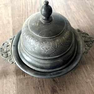 May include: Antique silver-toned butter dish with a domed lid and floral engravings. The dish has ornate handles and a round base. The lid is topped with a small, round knob. The butter dish is made of metal and sits on a wooden surface.