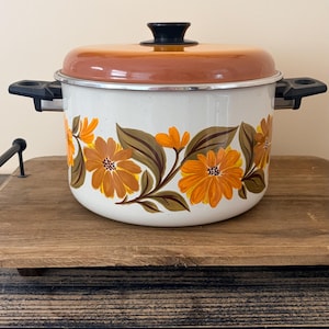 May include: White enamel pot with orange floral design and a brown lid. The pot has black handles and a black knob on the lid. A vintage kitchenware item.