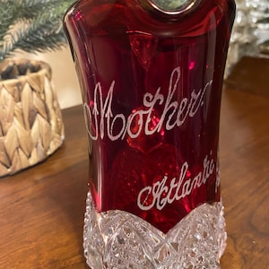 May include: A ruby red glass vase with the word "Mother" and "Atlantic" etched in a cursive font. The base of the vase has a cut crystal design. The vase is displayed on a wooden surface.
