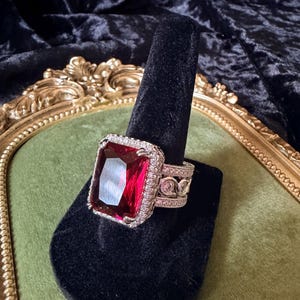 May include: A large, silver-toned ring with a rectangular, deep red gemstone. The gemstone is surrounded by a halo of smaller, clear stones. The ring band features intricate scrollwork and additional clear stones, creating a glamorous statement piece.
