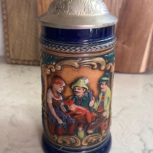 May include: A blue ceramic beer stein with a silver lid. The stein features a detailed relief scene of people and a fox. The relief is in shades of brown, orange, and green. The stein is a traditional German style.