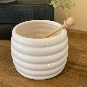 May include: White ceramic honey pot with a ridged design and a wooden honey dipper. The pot has a round shape and a wide opening. The honey dipper is light brown and has a small, round handle.
