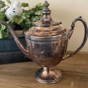 May include: A silver-colored teapot with a lid and curved handle. The teapot has a decorative design around the middle and sits on a pedestal base. The teapot is displayed on a wooden surface.
