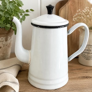 May include: A white enamel coffee pot with a black rim and a black knob on the lid. The pot features a curved spout, a side handle, and a cylindrical body. The white enamel finish shows minor signs of wear at the spout and base edges.