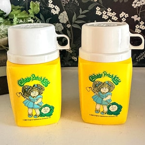 May include: Two yellow vintage Thermos bottles featuring the Cabbage Patch Kids logo. Each bottle has a white lid with a handle. The Cabbage Patch Kids logo includes a cartoon girl in a blue dress and a green cabbage.
