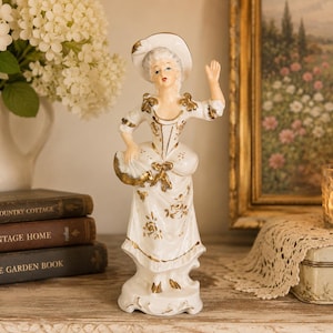 May include: A porcelain figurine of a person wearing a white dress and hat with gold accents. The figure stands on a base, holding a fan in one hand and raising the other hand. The attire features gold floral patterns and trim throughout the design.