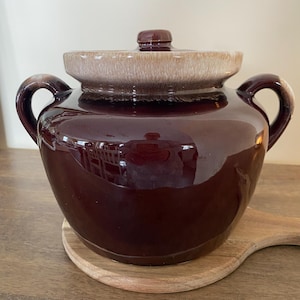 May include: A glossy, dark brown ceramic crock pot with a lid and two handles. The lid is two-toned, with a tan rim and a brown knob. The crock pot sits on a wooden cutting board.