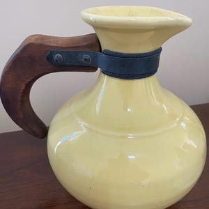 May include: A yellow ceramic pitcher with a dark wooden handle and a dark band around the neck. The pitcher has a rounded body and a spout. The handle is attached with metal hardware.